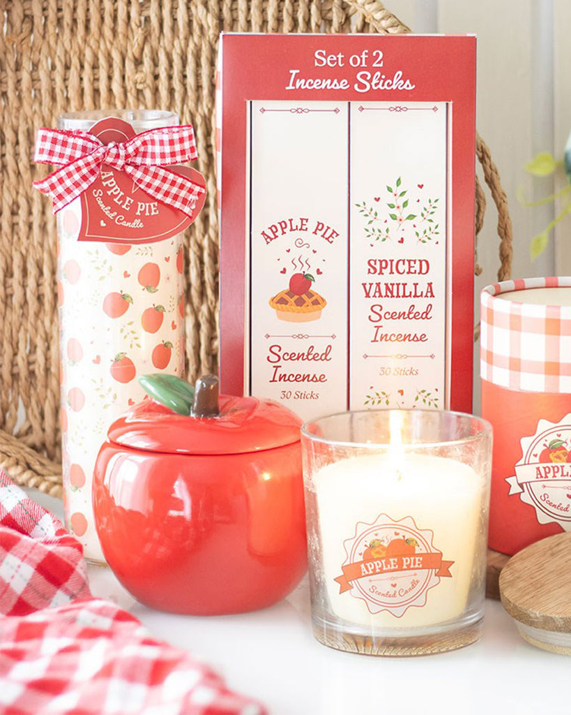 Lifestyle image featuring the apple-shaped candle jar as part of an apple-themed decor set, including a glass candle labeled 'Apple Pie' and a box of apple pie and spiced vanilla incense sticks.