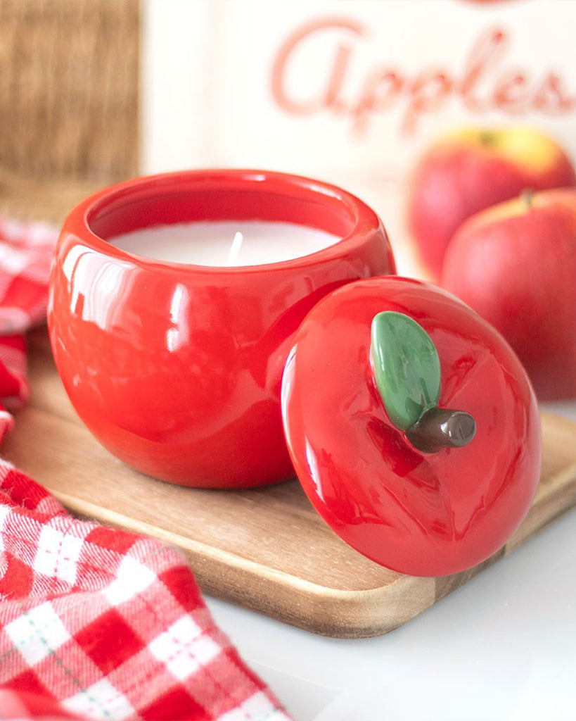 Ceramic apple-shaped candle jar with lid removed, showing a white wax candle inside. The red apple design includes a green leaf and brown stem on the lid, placed on a wooden tray with a red gingham cloth and apples in the background.