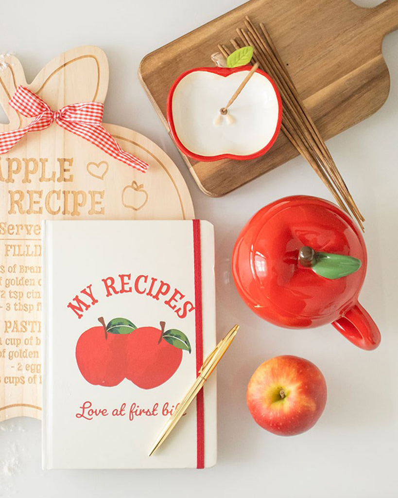 Flat lay image showing the apple mug with coordinating apple-themed accessories like a recipe book, incense holder, and a real apple.