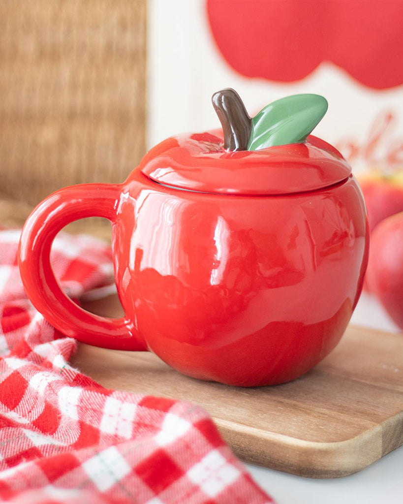 Apple-shaped ceramic mug with lid and handle, styled with a red checkered cloth and apple-themed decor in the background.