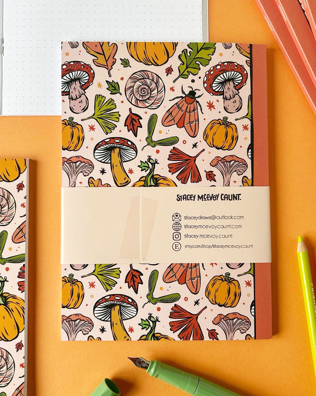 A stack of A5 notebooks featuring an autumnal pattern of pumpkins, mushrooms, leaves, and acorns on a soft peach background. The back of the notebook shows Stacey McEvoy Caunt’s branding with social media icons.