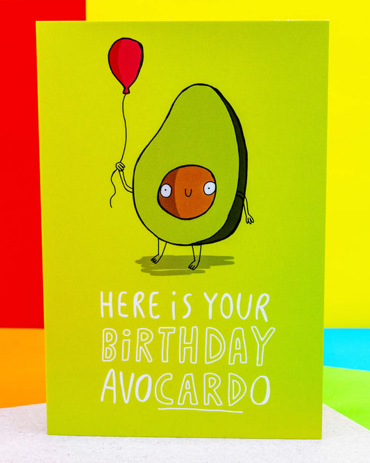 Bright green A6 greeting card by Rebel Romance featuring a cute illustrated avocado character by Katie Abey holding a red balloon. The card reads “Here is your birthday avocardo” in white text, with a quirky and playful style. Ideal for avocado lovers and pun fans, this fun birthday card is printed in the UK by a small alternative business celebrating all things extraordinary.
