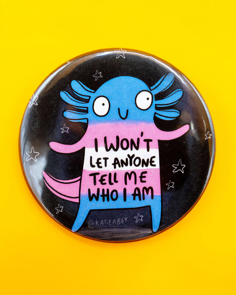 A flat lay of a Rebel Romance badge showing a colourful axolotl character in blue, pink and white with the message “I won’t let anyone tell me who I am”. Surrounded by star doodles on a black background, this Katie Abey design pops against a yellow backdrop.