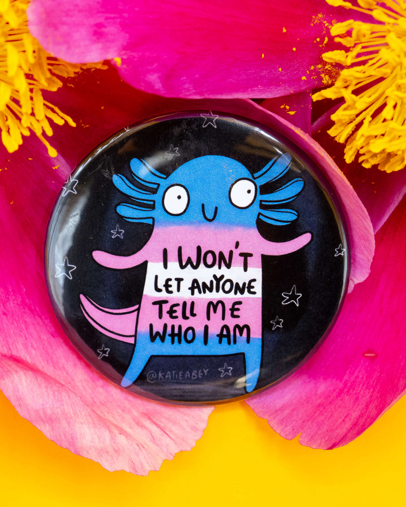 Rebel Romance round badge displayed on vivid pink and yellow flower petals. The design features an axolotl in trans flag colours with the words “I won’t let anyone tell me who I am”. A bold celebration of identity, illustrated by Katie Abey.
