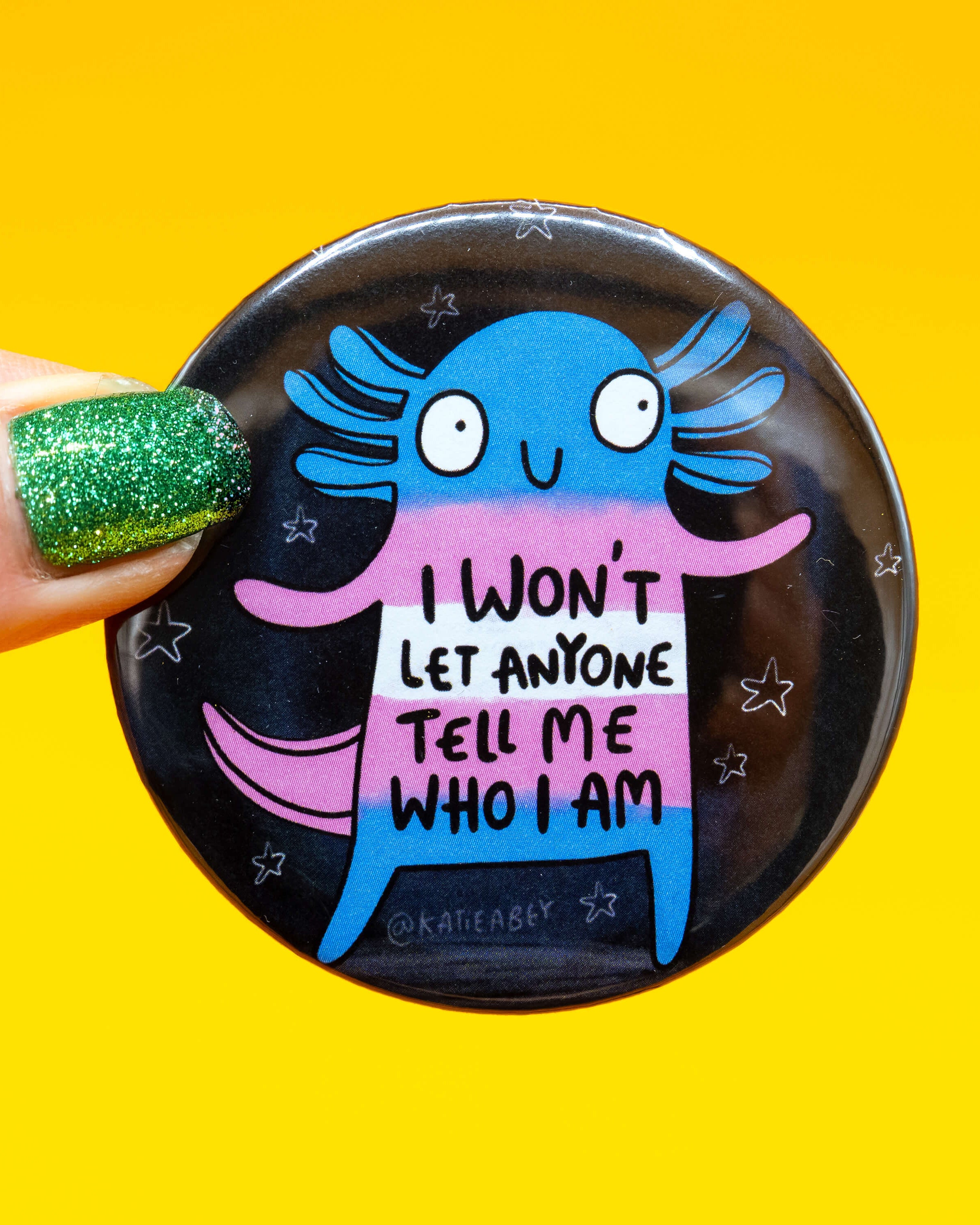 Close-up of a hand holding a Rebel Romance round badge featuring a bright axolotl in the trans pride flag colours—blue, pink and white—against a starry black background. Text on the badge reads “I won’t let anyone tell me who I am”. Designed by Katie Abey. Set against a vibrant yellow background.