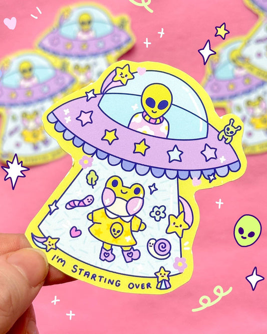Glossy pastel UFO sticker from Rebel Romance featuring a kawaii yellow alien abducting a frog character in a cute outfit with the words "I'm Starting Over." Surrounded by stars, worms, and snails, this fun vinyl sticker is perfect for scrapbooking, journaling, or decorating your laptop or notebook. Designed by Sun Puff Studios. A waterproof and durable sticker for fans of space, aliens, and pastel aesthetics.