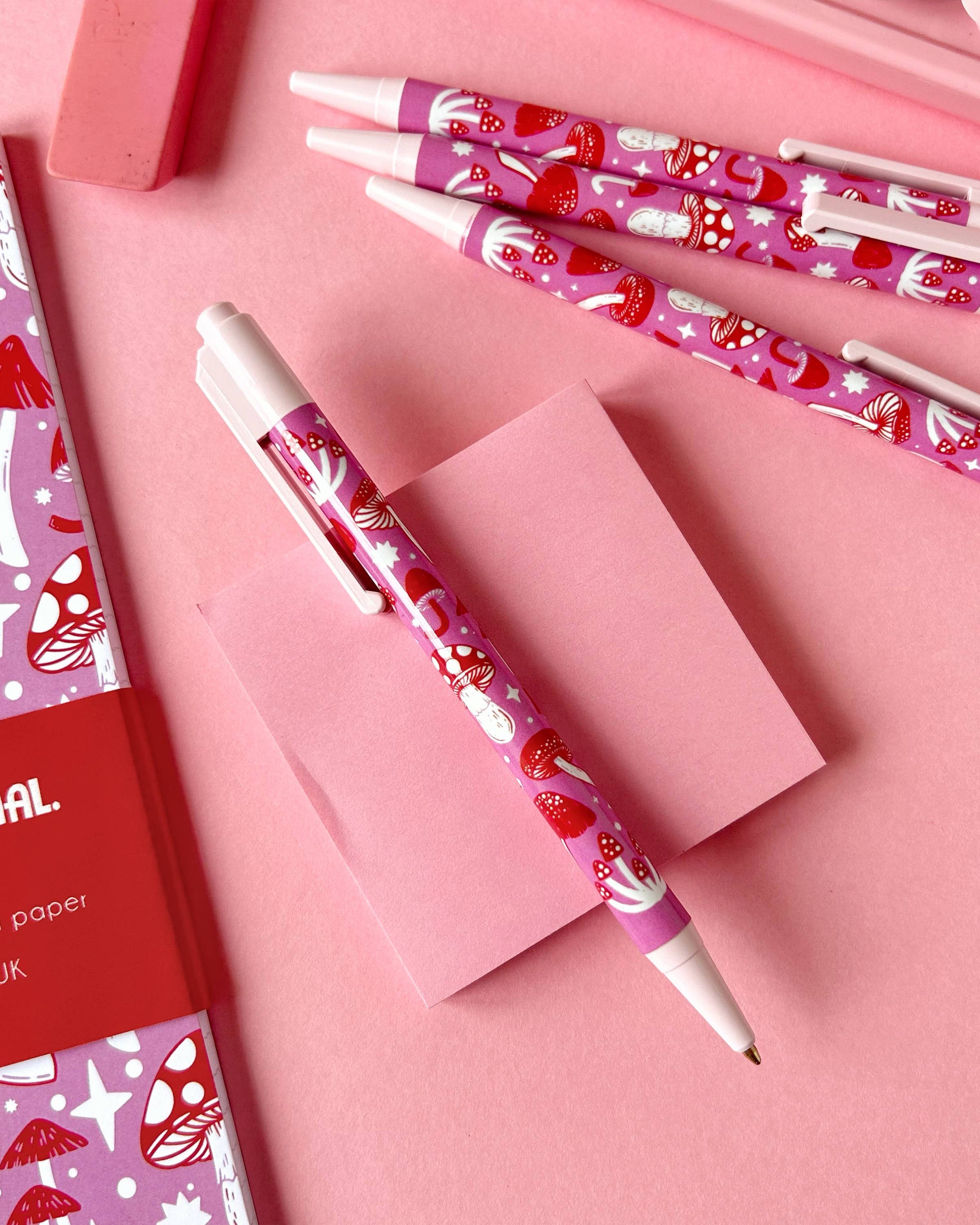 Flat lay of a pink and red mushroom-patterned pen on a pink sticky note, surrounded by matching pens, pink stationery, and a notebook. Designed by Stacey McEvoy Caunt for Rebel Romance, a small UK business celebrating alternative stationery and aesthetics.