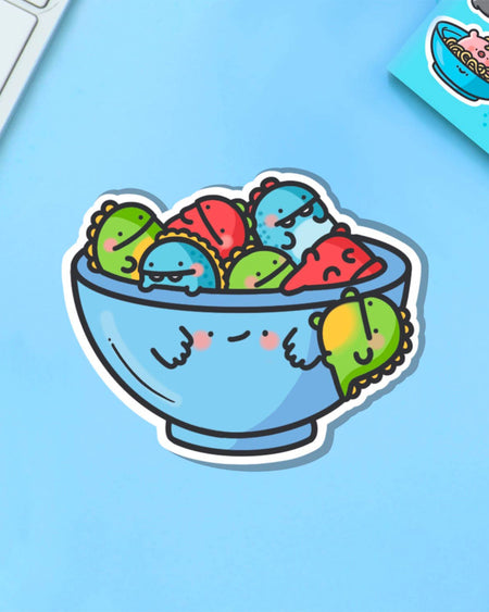 Close-up of Toastedink's Bowl of Dinosaurs vinyl sticker on a blue background, showing detailed kawaii-style dinosaurs in a happy bowl. Fun, waterproof and perfect for laptops or journals. Stocked by UK brand Rebel Romance.