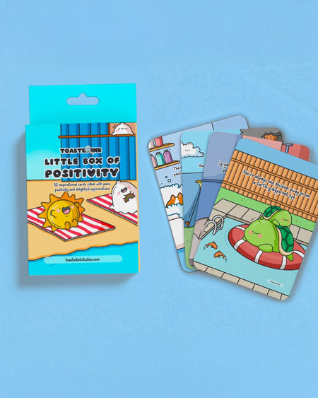 A blue box titled “Little Box of Positivity” surrounded by several colourful Toastedink affirmation cards with punny, illustrated designs.