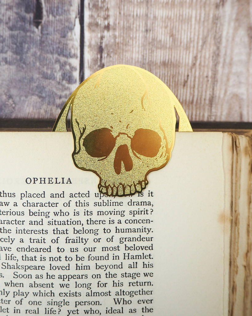 Gold brass skull bookmark peeking from the top of a Hamlet book page. A Literary Emporium creation available at Rebel Romance, a UK business known for gothic gifts and alternative style.