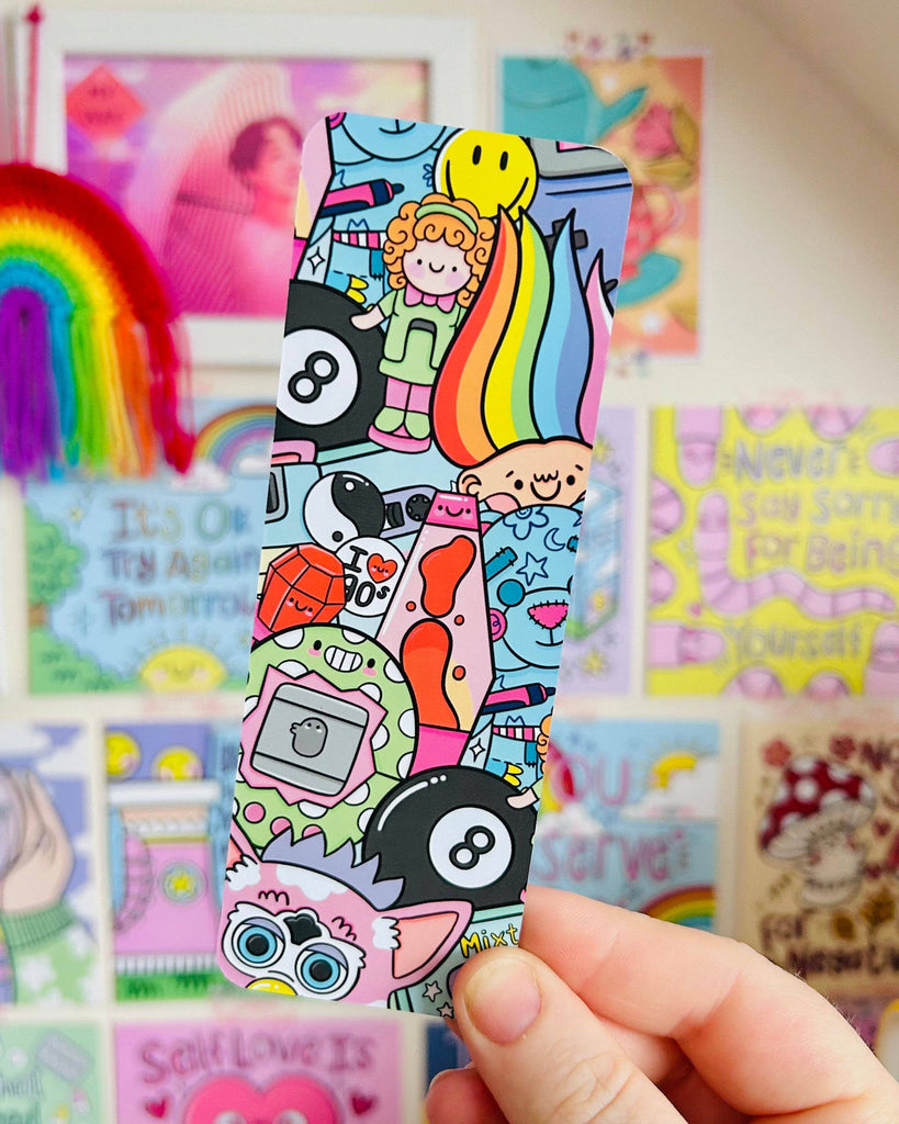 Hand holding the 90s Nostalgia Bookmark by Kelly Lou, surrounded by vibrant, retro-inspired wall art. This velvet-laminated bookmark from Rebel Romance features fun 90s throwbacks including a rainbow troll, Furby, mixtape, Tamagotchi, and smiley face. A colourful, quirky stationery gift from a UK small business celebrating alternative aesthetics.
