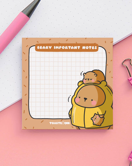 Square sticky note pad by Toastedink, sold at Rebel Romance, titled "Beary Important Notes" featuring a bear in a yellow hoodie with cubs. Set on a pastel pink background with a pen and paperclip.