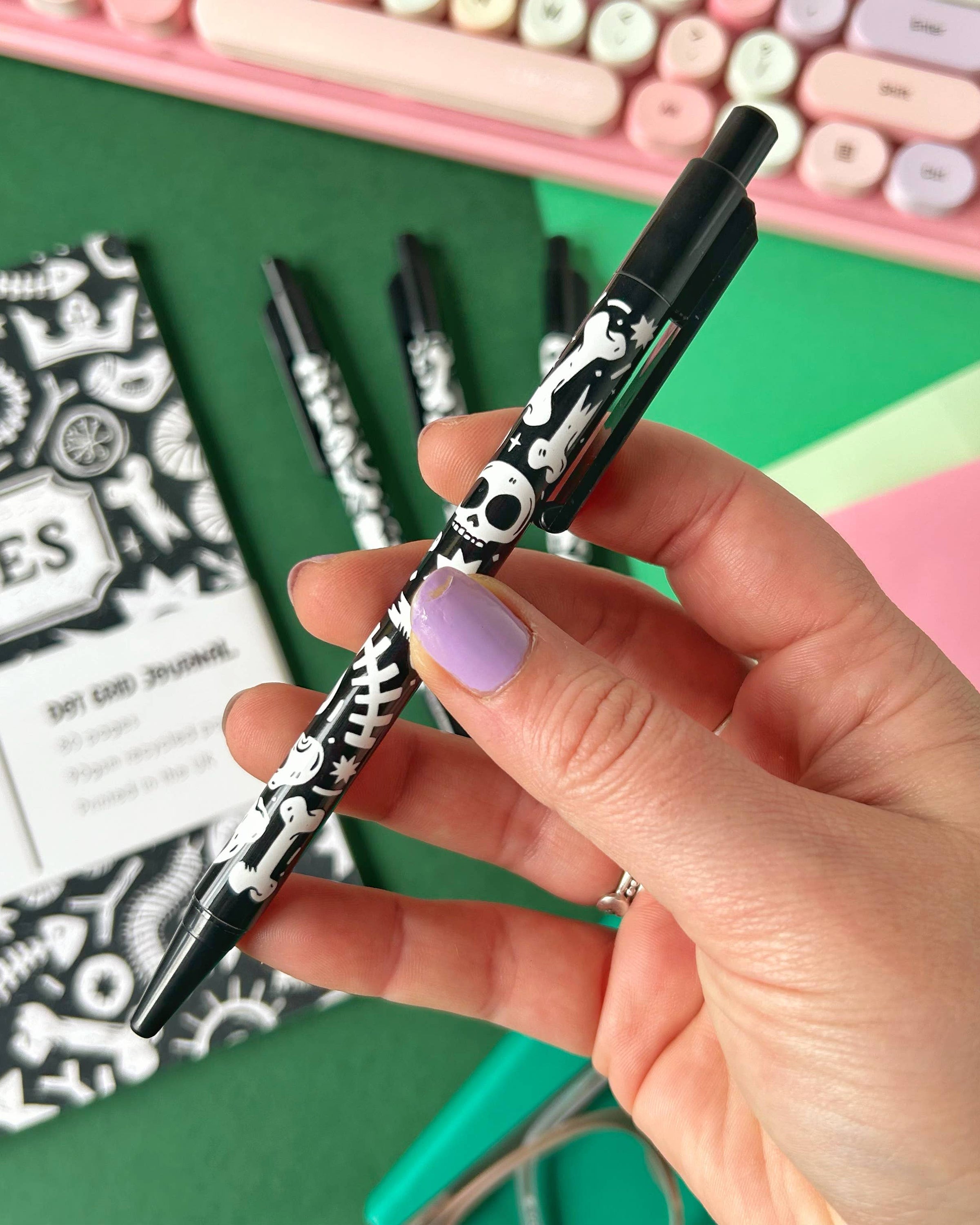 Close-up of a hand holding a Rebel Romance ballpoint pen decorated with Stacey McEvoy Caunt’s skull and bones pattern, perfect for lovers of dark and quirky stationery. Background features other pens, a notebook, and sticky notes.