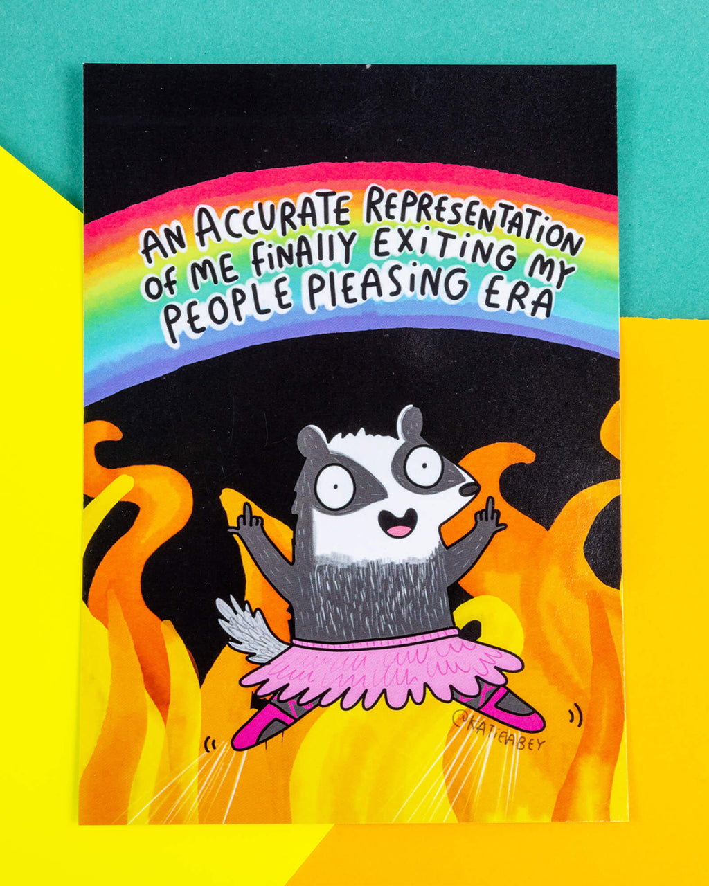 A postcard of a badger in a pink ballet outfit giving two fingers up on a black and fiery illustration with a rainbow above them saying 'An accurate representation of me finally exiting my people pleasing era'. Illustrated by Katie Abey