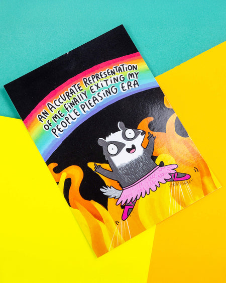 A postcard of a badger in a pink ballet outfit giving two fingers up on a black and fiery illustration with a rainbow above them saying 'An accurate representation of me finally exiting my people pleasing era'. Illustrated by Katie Abey.