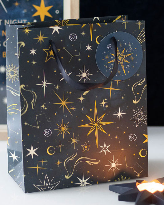 Medium Starry Night gift bag styled with glowing candles and a dark blue backdrop, showcasing its golden star design and ribbon handles.