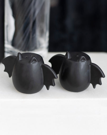 A set of black ceramic bat wing salt and pepper shakers sitting on a white surface with a dark background. Their quirky bat wing shape adds a gothic touch to any dining table.