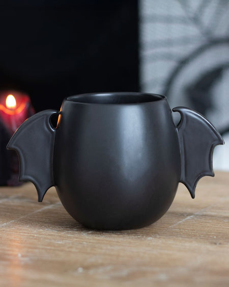 Styled lifestyle photo of the Bat Wing Mug on a wooden table with a black candle in the background. The bat wing handles and matte black ceramic make it an eye-catching gothic gift.