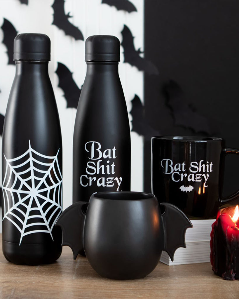 A black bat-themed homeware set featuring two water bottles (one with a spiderweb, one reading "Bat Shit Crazy"), a black mug with the same slogan, and a round bat wing mug with sculpted wing handles. Perfect for gothic and Halloween lovers.