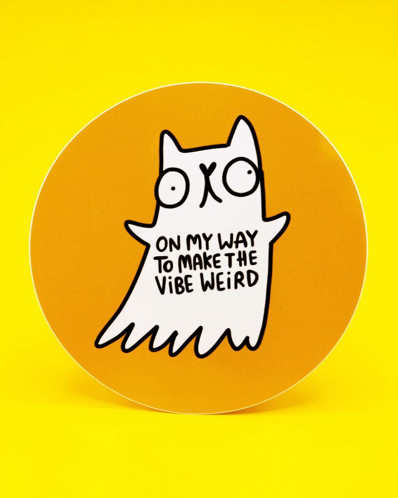 A round vinyl sticker featuring a quirky white ghost cat illustration by Katie Abey on an orange background. The ghost cat has wide eyes and the text reads On My Way To Make The Vibe Weird. Premium vinyl sticker, 10 cm diameter, durable and wipe clean. Perfect for laptops, notebooks, water bottles, and gifts for people who love cute and weird art.
