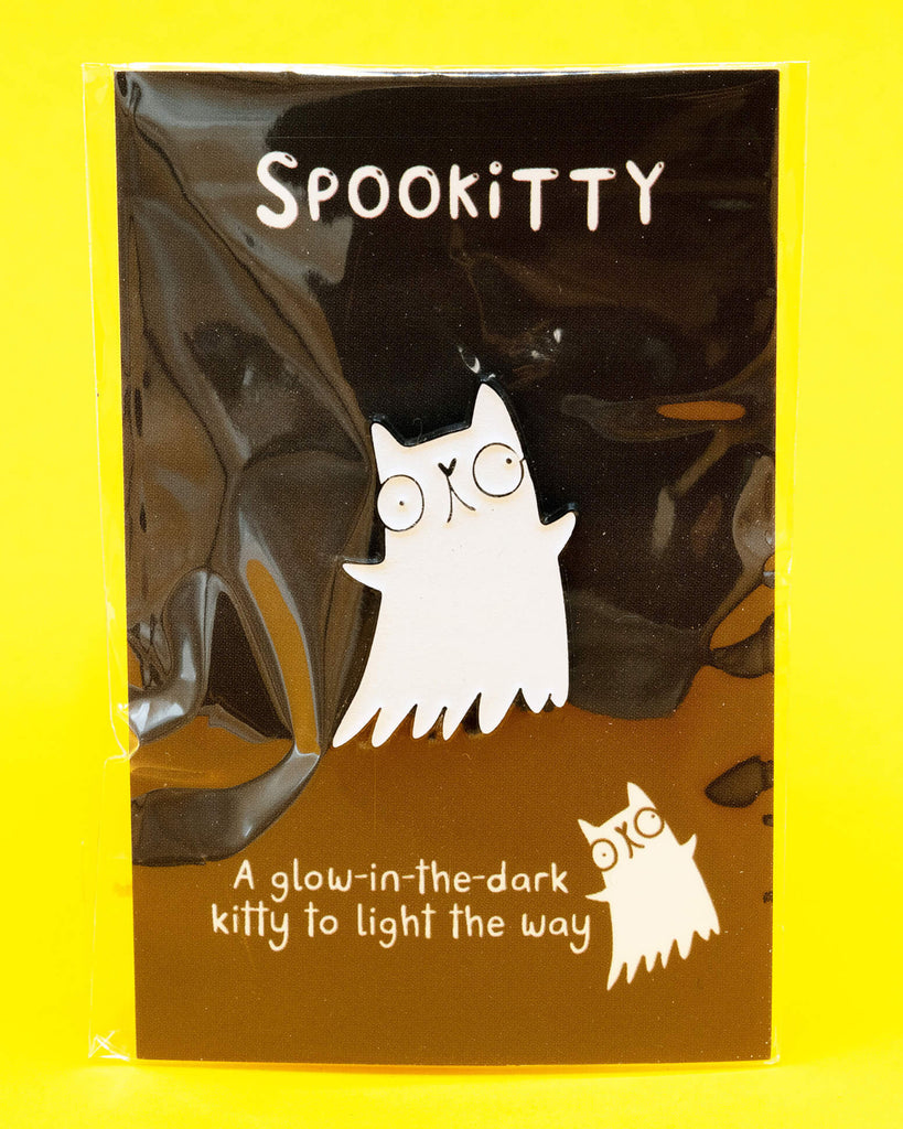 A Katie Abey Spookitty glow in the dark glitter enamel pin badge shown on its black backing card inside clear packaging against a bright yellow background. The ghost cat enamel pin has a white glitter finish and a bold black outline. Fun spooky themed accessory for bags, denim jackets, and pin collections.