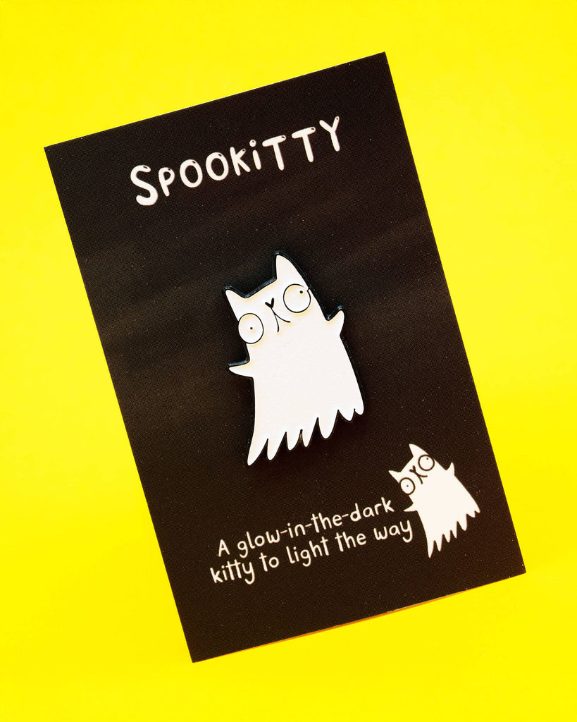 A Katie Abey Spookitty glow in the dark glitter enamel pin badge displayed on a black backing card against a bright yellow background. The white ghost cat design features big round eyes, a black outline, and a wavy ghost tail. Cute spooky themed enamel pin perfect for jackets, bags, and accessories.