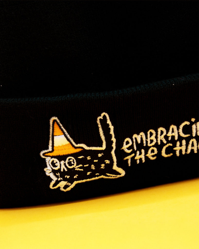 Close up of the embroidered design on the black Embracing The Chaos Cat Beanie Hat by Katie Abey. The detail shows a cute chaotic cat wearing a candy corn coloured hat with the words Embracing The Chaos. Ethically made quirky beanie perfect for alternative fashion and Katie Abey fans.