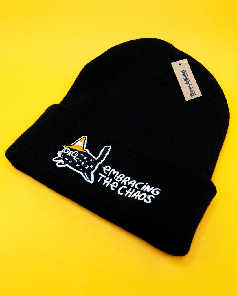 A black Embracing The Chaos Cat Beanie Hat by Katie Abey shown on a bright yellow background. The beanie features an embroidered quirky black cat wearing a candy corn coloured hat with the text Embracing The Chaos stitched in white. Cosy ethically made winter hat perfect for quirky fashion lovers and fans of Katie Abey.