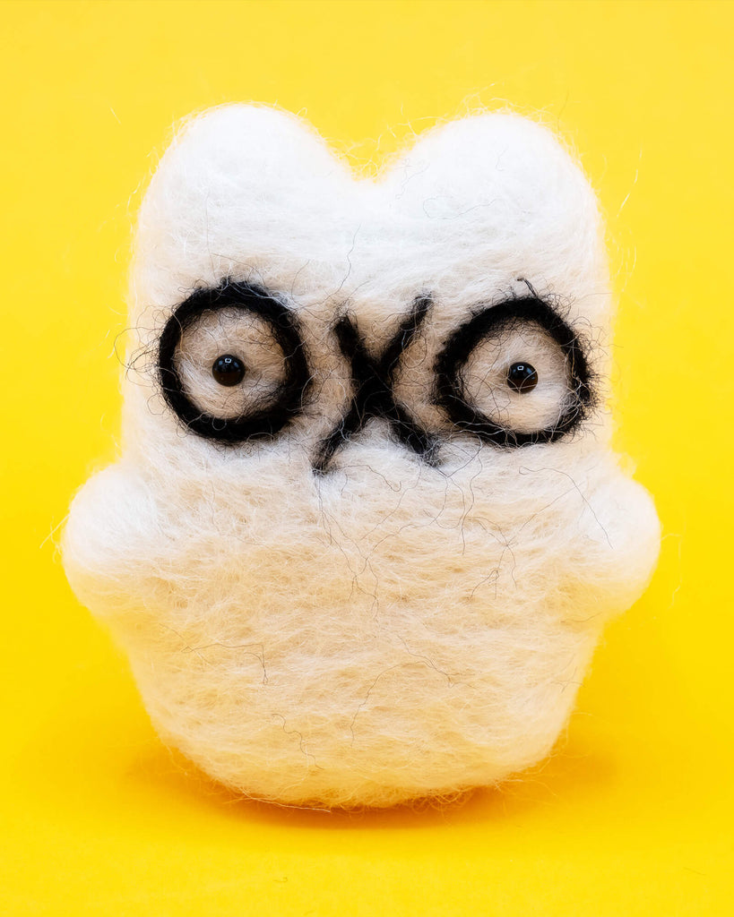 A handmade needle felted ghost cat by Katie Abey x Ghost Of The Month shown on a bright yellow background. The white wool ghost cat has big black outlined eyes and a cute quirky expression. Felted by hand, approx 8cm by 5cm, this decorative collectible is perfect for fans of weird cute art and handmade gifts.