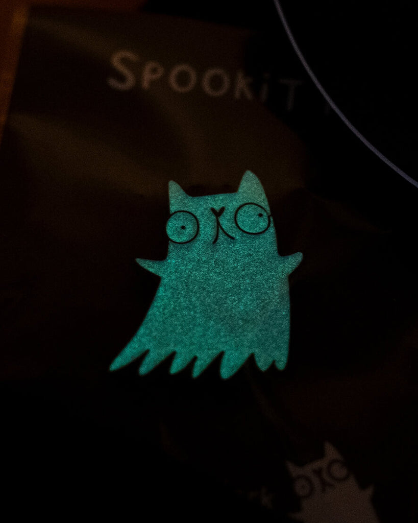 A Katie Abey Spookitty glow in the dark glitter enamel pin badge glowing bright green in the dark. The ghost cat shape is clearly visible with round eyes and wavy edges. A playful spooky themed enamel pin that glows at night and stands out on jackets, bags, and accessories.