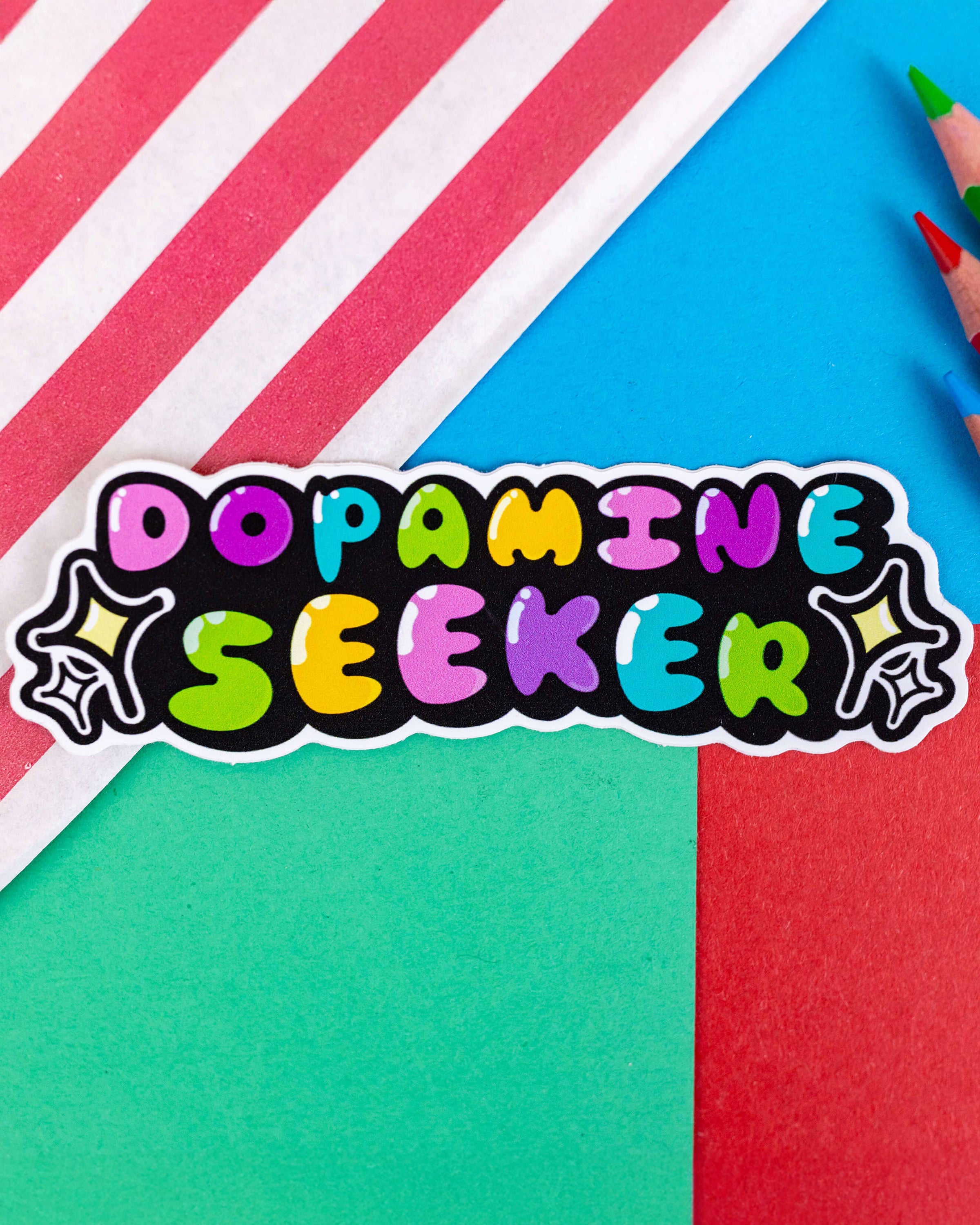 A colourful dopamine-themed vinyl sticker reading “Dopamine Seeker” in bold, bubbly rainbow letters with white highlights on a black background. The custom-shaped sticker includes star sparkle accents and sits against a vibrant background of red, blue, green, and pink-striped paper. Designed by Innabox and sold by Rebel Romance, a UK small business celebrating alternative style.