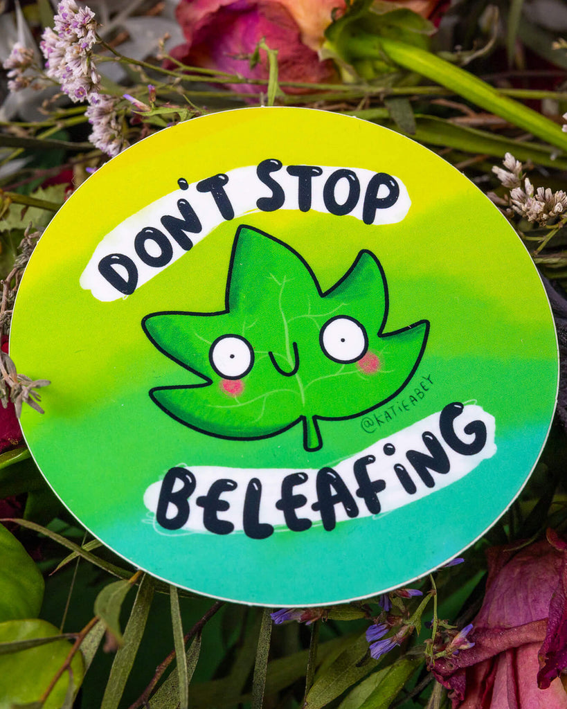 A round green vinyl sticker designed by Katie Abey, available at Rebel Romance. The sticker features a wide-eyed, smiling cartoon leaf with rosy cheeks, surrounded by the pun “Don’t Stop Beleafing” in playful black and white text. A cute and quirky design, perfect for plant lovers and positivity fans.