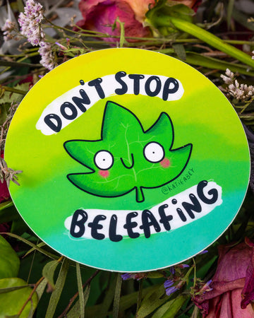 A round green vinyl sticker designed by Katie Abey, available at Rebel Romance. The sticker features a wide-eyed, smiling cartoon leaf with rosy cheeks, surrounded by the pun “Don’t Stop Beleafing” in playful black and white text. A cute and quirky design, perfect for plant lovers and positivity fans.