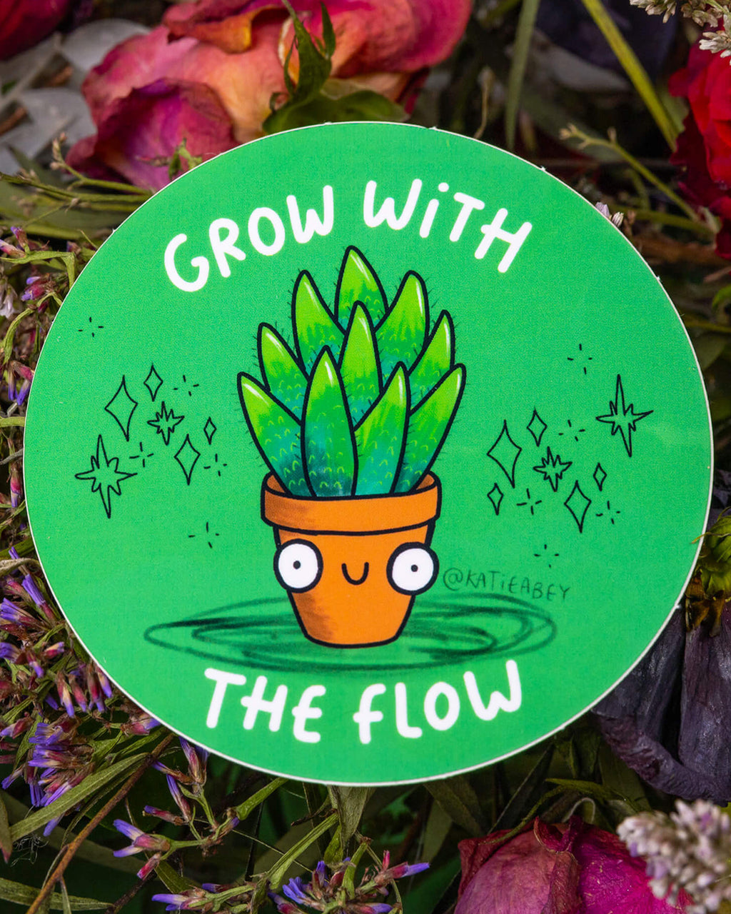 A round green vinyl sticker by Katie Abey, available from Rebel Romance. The sticker features a smiling cartoon plant in a terracotta pot with wide eyes and leafy head, surrounded by sparkles and the phrase “Grow With The Flow” in white text. A cheerful and encouraging design, perfect for personalising notebooks, bottles, and laptops.