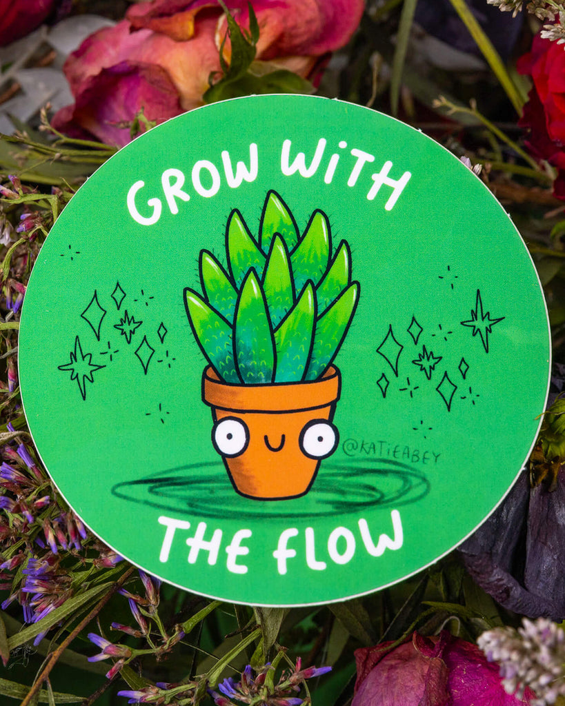 A round green vinyl sticker by Katie Abey, available from Rebel Romance. The sticker features a smiling cartoon plant in a terracotta pot with wide eyes and leafy head, surrounded by sparkles and the phrase “Grow With The Flow” in white text. A cheerful and encouraging design, perfect for personalising notebooks, bottles, and laptops.