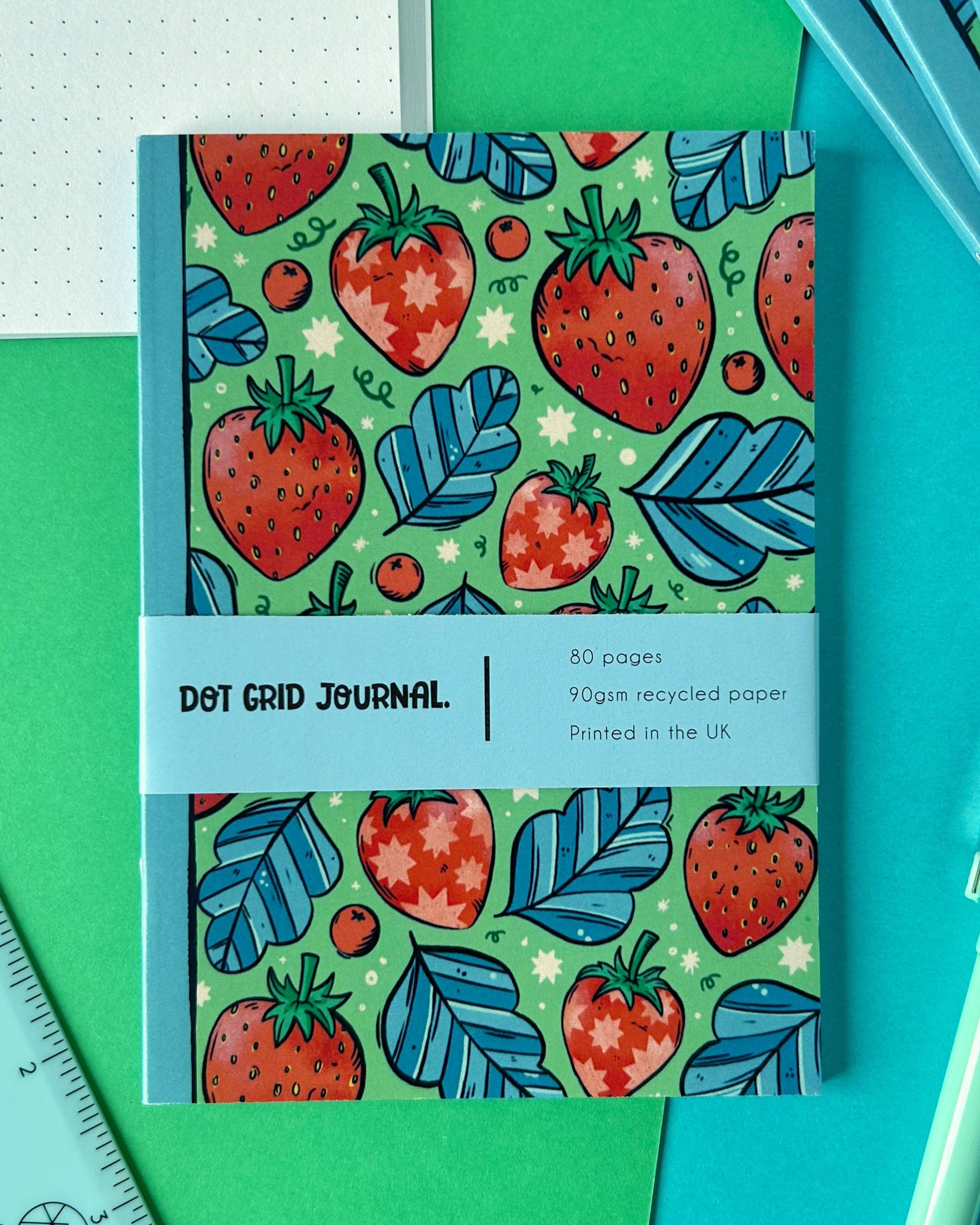 A colourful A6 dot grid journal by Rebel Romance featuring a bright strawberry and blue leaf pattern on a green background. The cover wrap reads “Dot Grid Journal – 80 pages, 90gsm recycled paper, Printed in the UK.” The notebook is styled on green and blue paper with a dotted journal page in the background.