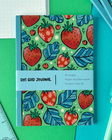 A colourful A6 dot grid journal by Rebel Romance featuring a bright strawberry and blue leaf pattern on a green background. The cover wrap reads “Dot Grid Journal – 80 pages, 90gsm recycled paper, Printed in the UK.” The notebook is styled on green and blue paper with a dotted journal page in the background.