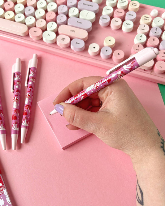 A hand with lilac nails writing with the pink mushroom pen by Rebel Romance, with a pastel keyboard and pink notepad in the background. The pen features a cute red and pink mushroom print designed by Stacey McEvoy Caunt.