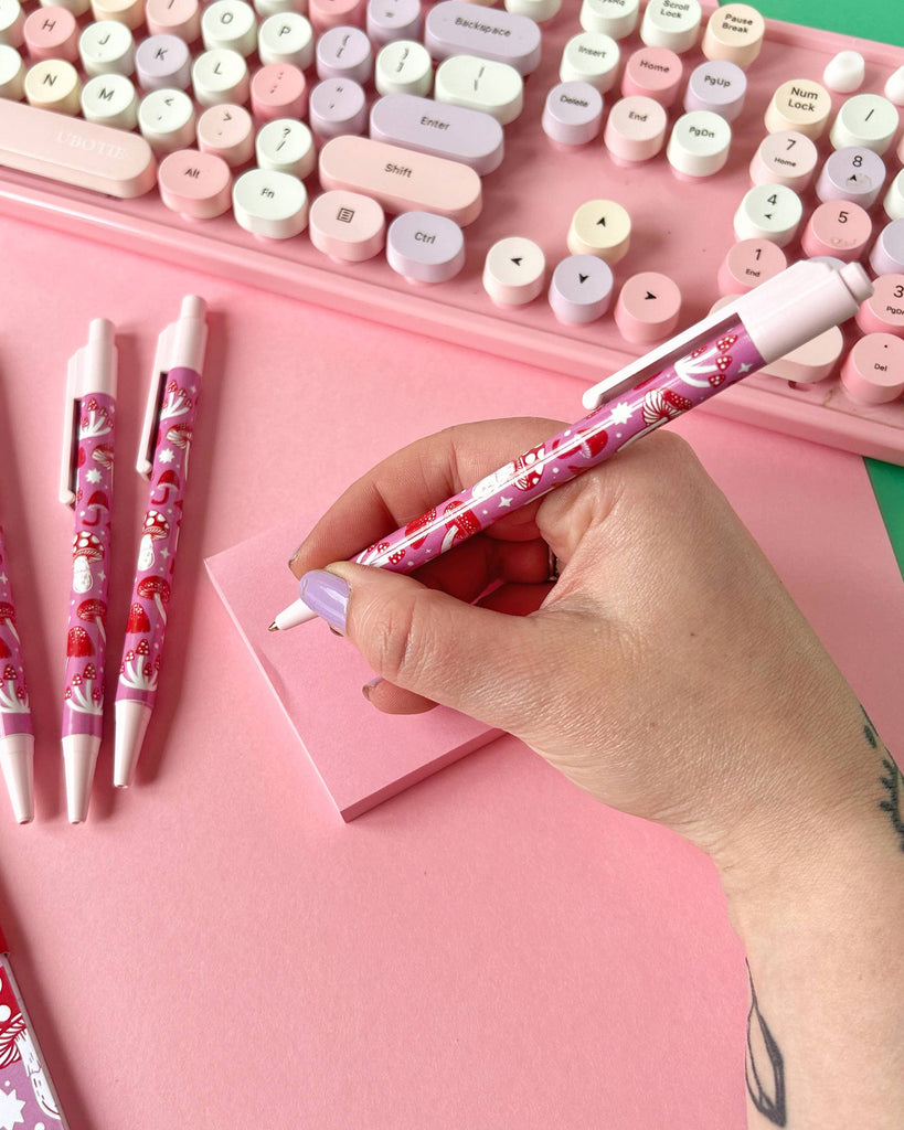 A hand with lilac nails writing with the pink mushroom pen by Rebel Romance, with a pastel keyboard and pink notepad in the background. The pen features a cute red and pink mushroom print designed by Stacey McEvoy Caunt.