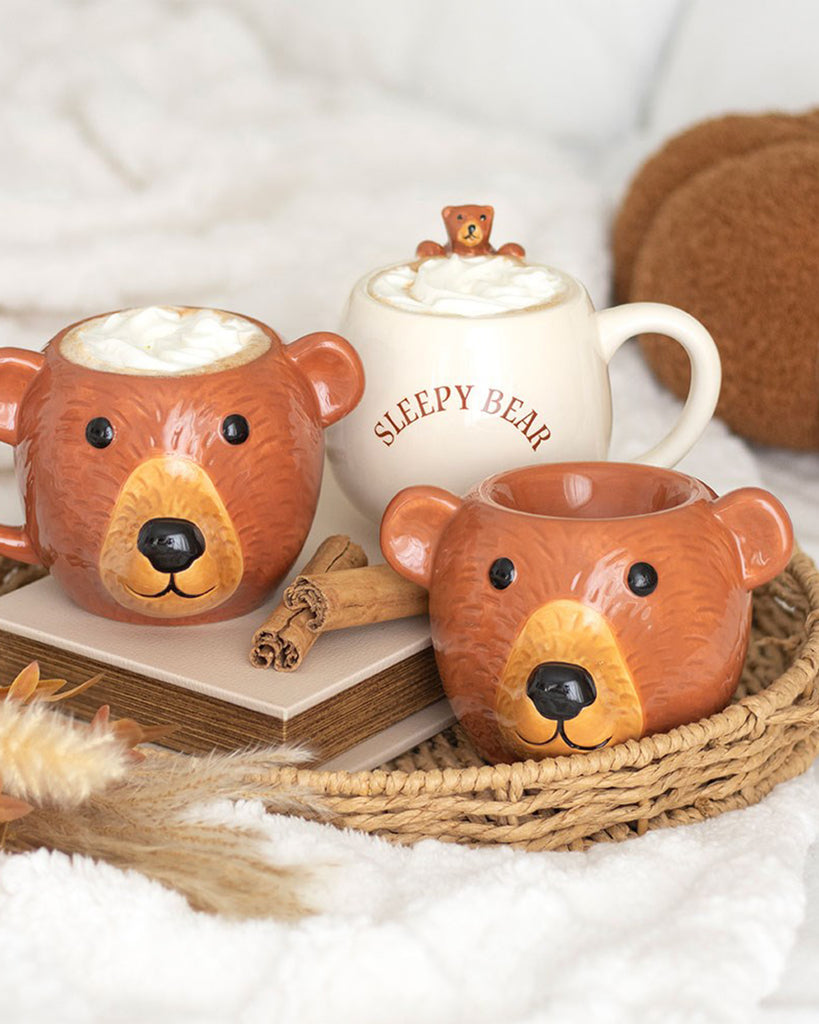 Cosy flatlay of bear-shaped mugs and a cream ceramic "Sleepy Bear" mug with whipped cream on top, displayed on a fluffy blanket with cinnamon sticks, books and a woven tray. Ideal for autumn or winter vibes, this cute homeware setup features the Rebel Romance Bear Oil Burner adding a whimsical touch to the scene.