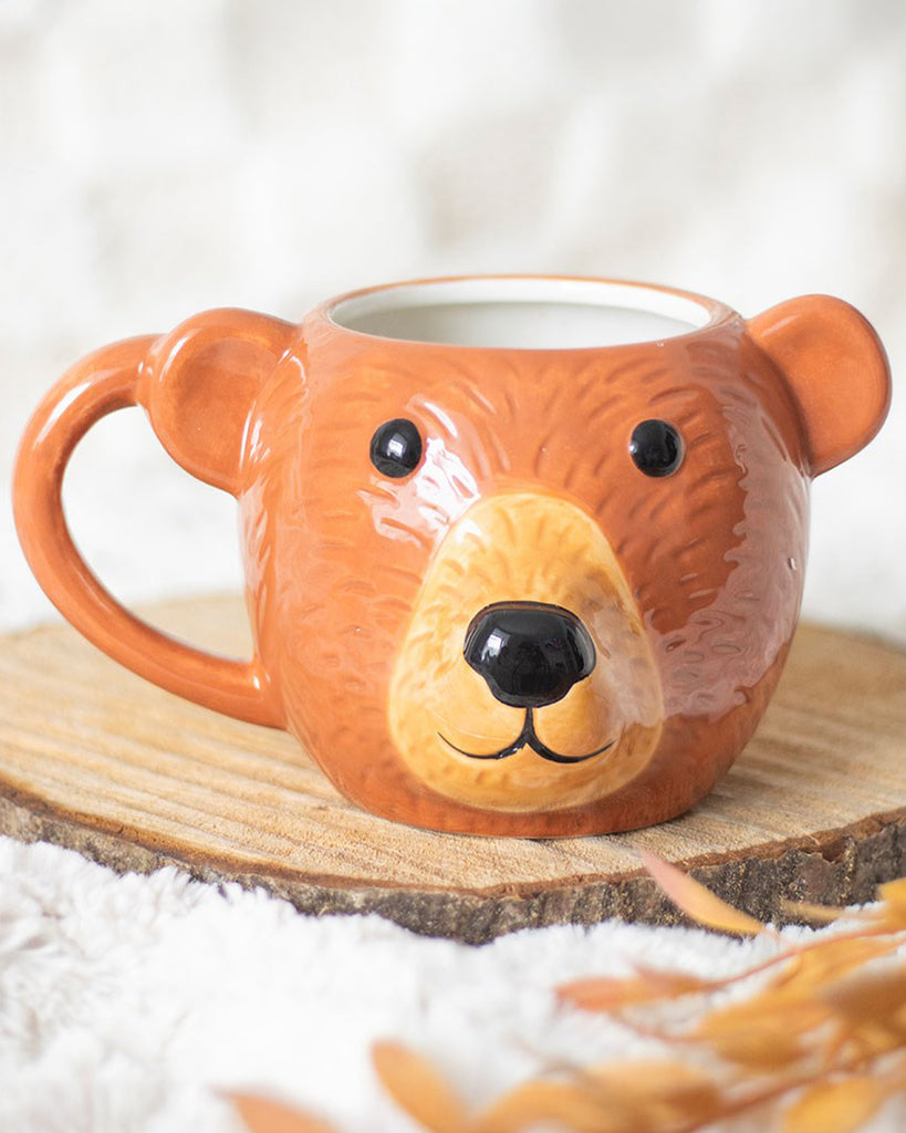 Close-up of the Rebel Romance Bear Shaped Mug sitting on a wooden coaster with cosy white bedding and autumn leaves, highlighting its cute bear face and orange ceramic glaze. A unique gift idea for tea, coffee or hot chocolate lovers.