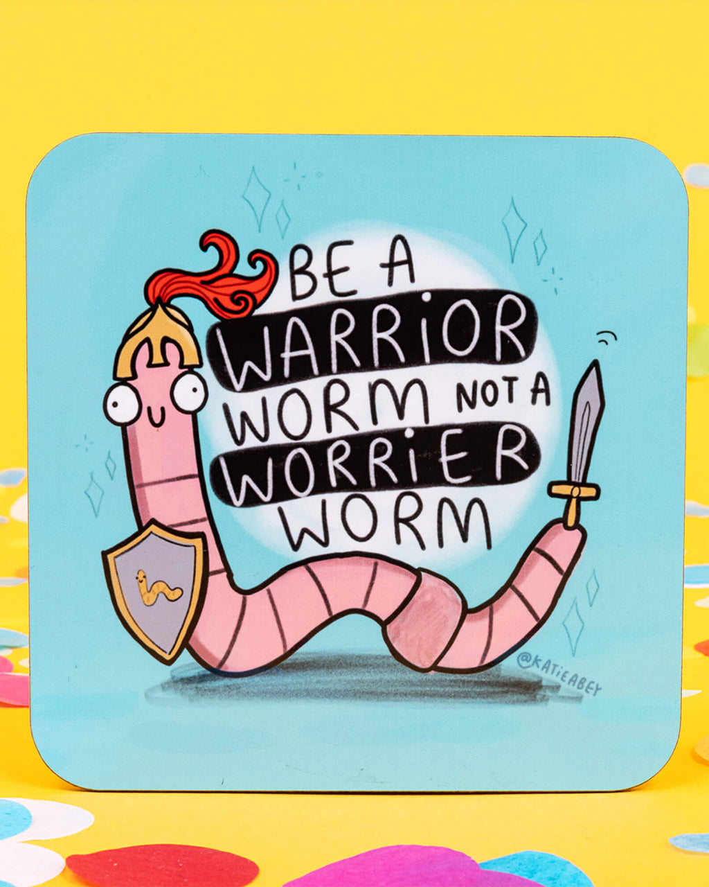 A square coaster with a blue background featuring a pink cartoon worm wearing a helmet and holding a sword and shield. The text says: “Be a warrior worm not a worrier worm.” Designed by Katie Abey. Coaster measures approximately 9cm square with rounded corners. A motivational and quirky design ideal for desks, coffee tables, and gifting.