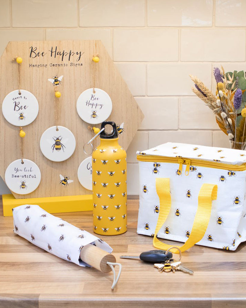 A cheerful collection of bee-themed gifts and homeware from Rebel Romance, a small UK business embracing alternative style. The Bee Metal Water Bottle is front and centre, featuring a vibrant yellow background with an all-over bee print, a black screw-top lid, and a handy clip. Surrounding it are matching bee-themed items, including a cool bag, umbrella, and ceramic hanging signs, all part of the Bee Happy collection.