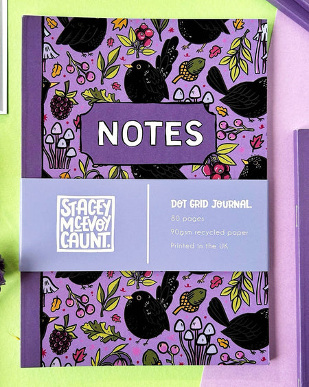 A close-up of the Blackbird Notebook, showing a vibrant purple cover with illustrations of blackbirds, mushrooms, berries, and leaves. The cover has a label reading “NOTES” with a lavender band listing product details: “Dot Grid Journal, 80 pages, 90gsm recycled paper, Printed in the UK.”