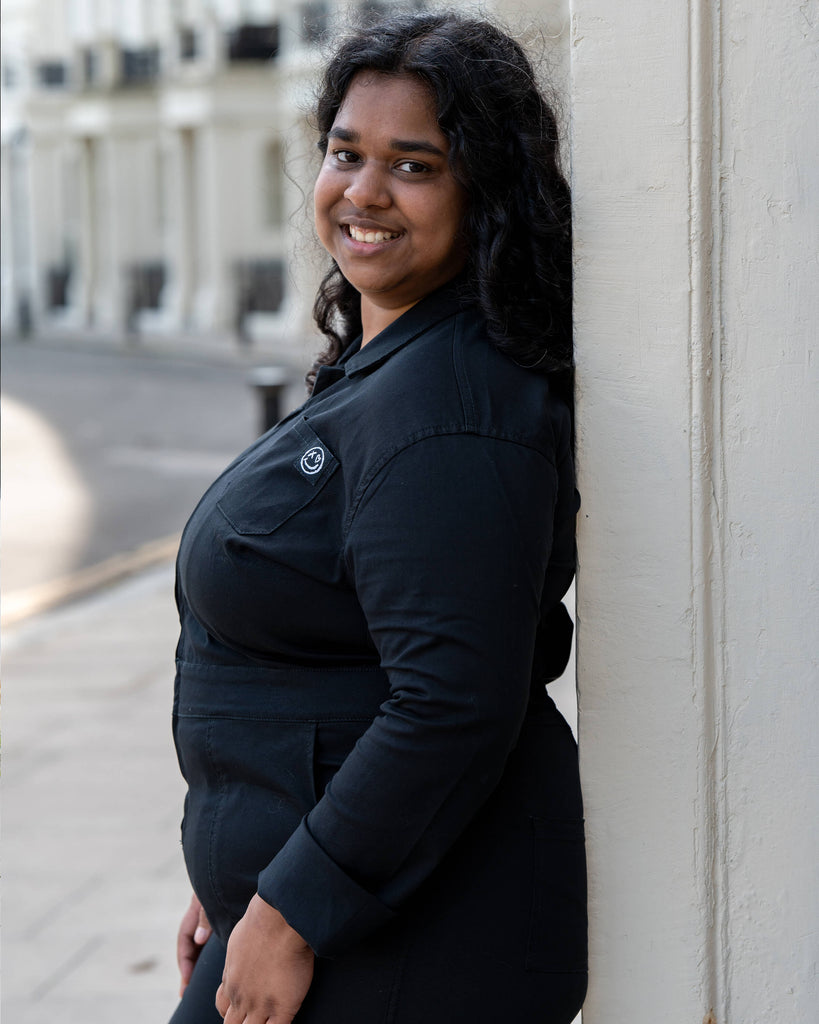 Vidhya leans against a cream pillar in the Rebel Romance Jet Black Boiler Suit, smiling with relaxed confidence. This sleek all-in-one is made from stretch cotton twill and designed for everyday edge and comfort.