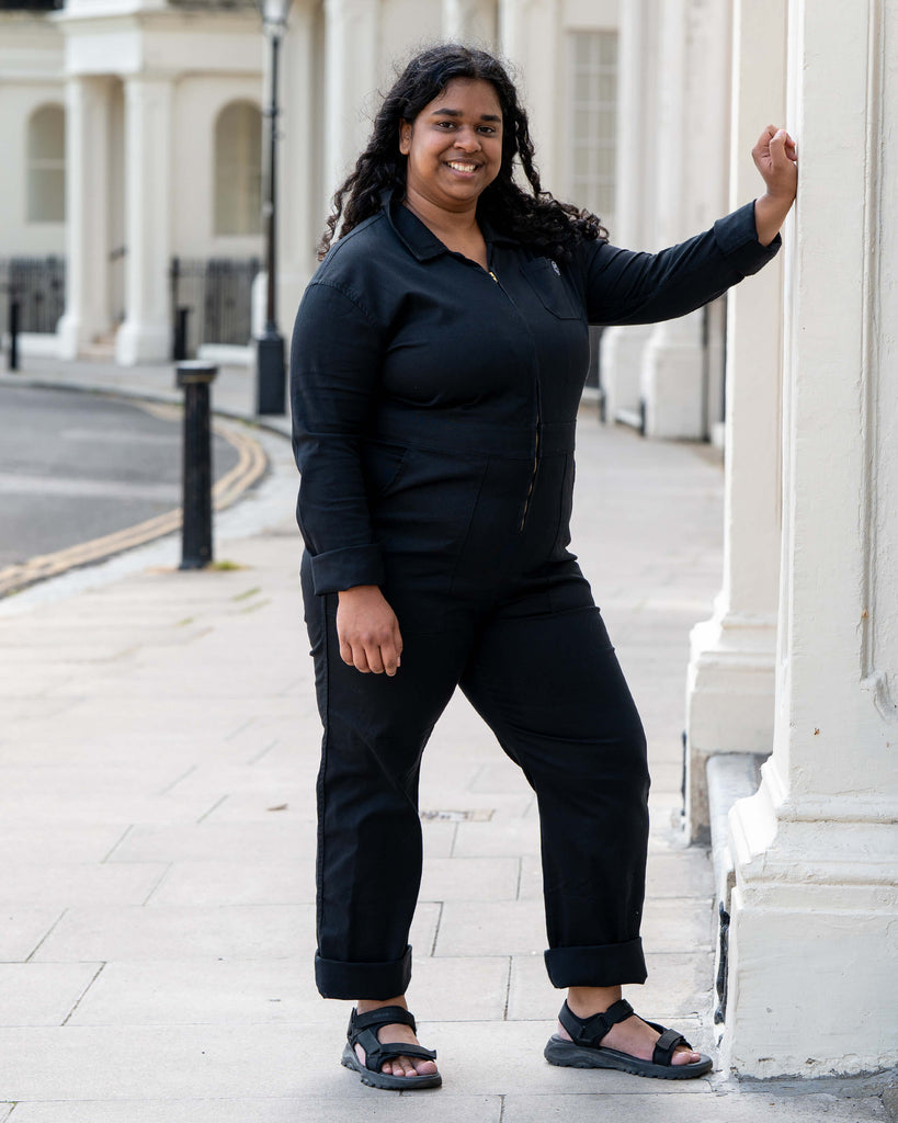 Vidhya stands confidently with one hand on a column, modelling the Jet Black Boiler Suit by Rebel Romance. An inclusive, ethical fashion piece that’s bold, comfy, and totally wearable.
