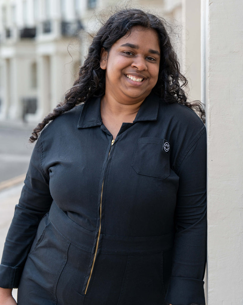 Vidhya smiles warmly while leaning against a cream building in the Jet Black Boiler Suit by Rebel Romance. This versatile all-in-one balances comfort, structure, and alternative style effortlessly.
