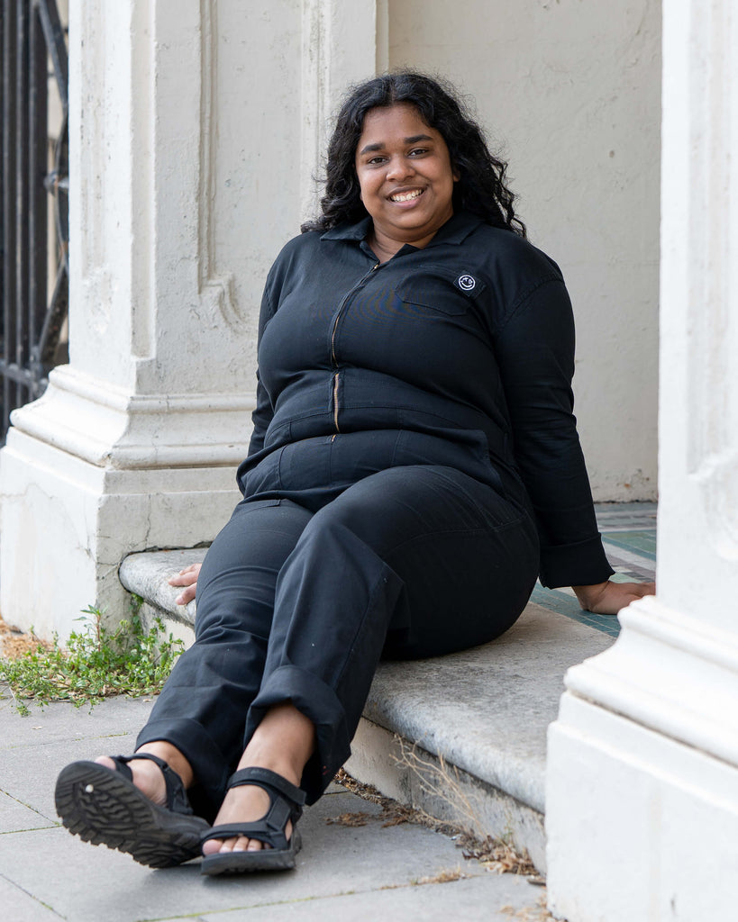 Model Vidhya sits casually on a stone step in the Jet Black Boiler Suit by Rebel Romance, showing off its easy fit and streetwear appeal. Perfect for slow fashion lovers who like a practical but stylish edge.