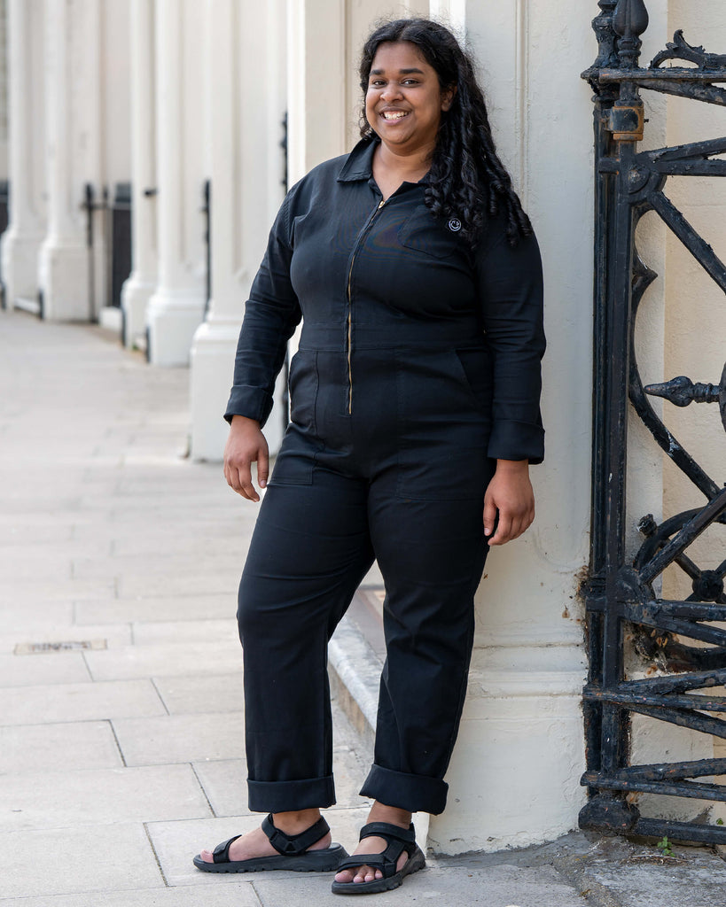 Vidhya poses by a cream building in the Rebel Romance Jet Black Boiler Suit, smiling brightly. A confident and comfy fit that brings alternative fashion vibes to everyday dressing.