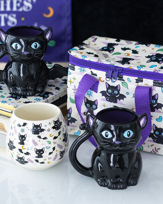 Cute and Creepy Black Cat Mug by Rebel Romance displayed with matching gothic cat print accessories including a cooler bag, notebook, and cauldron-style mug. This glossy ceramic mug features bright blue eyes, a curled tail handle, and a deep purple interior. A spooky and stylish gift idea for Halloween lovers and fans of alternative aesthetics.